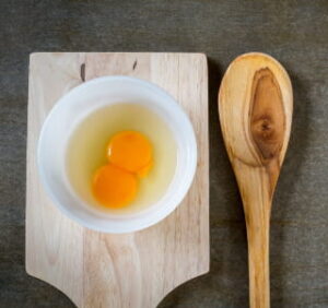 bowl on a cutting board with a double-yolked egg inside and a wooden spoon next to them